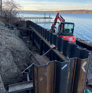 Excavator installing steel sheet piling along a rocky shoreline